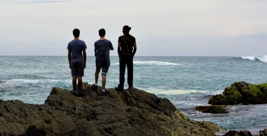 Three young people standing together on a rocky headland, watching the swell